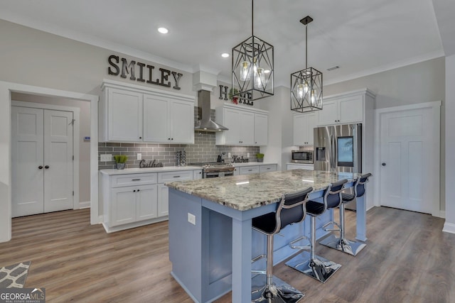 kitchen with appliances with stainless steel finishes, wall chimney exhaust hood, white cabinets, a center island, and hanging light fixtures