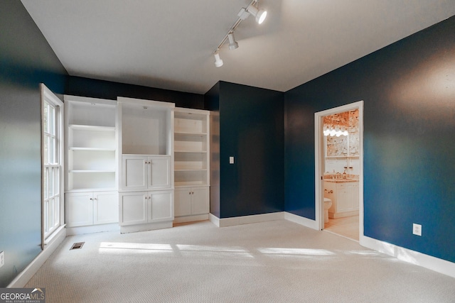 unfurnished bedroom featuring multiple windows, track lighting, connected bathroom, and light carpet