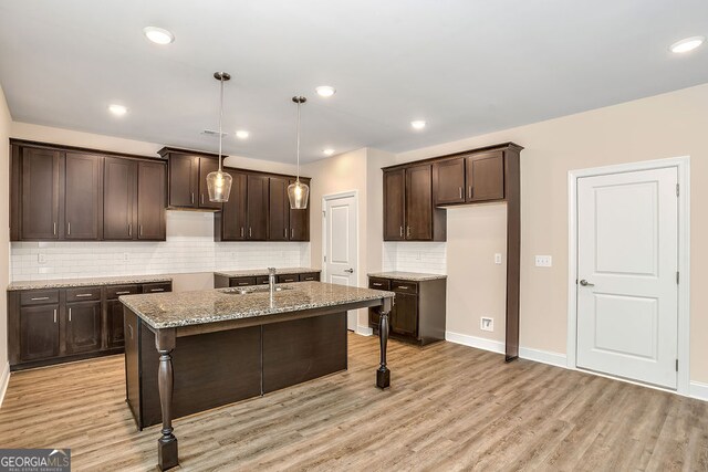 kitchen featuring a center island with sink, hanging light fixtures, decorative backsplash, light hardwood / wood-style floors, and light stone counters