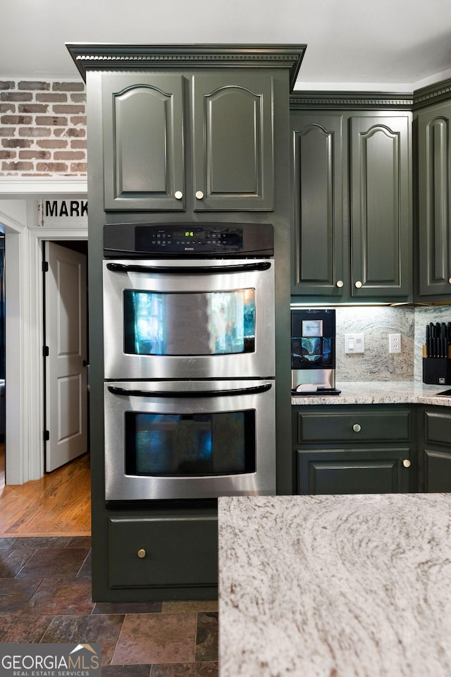 kitchen with stainless steel double oven, backsplash, light stone counters, and green cabinetry