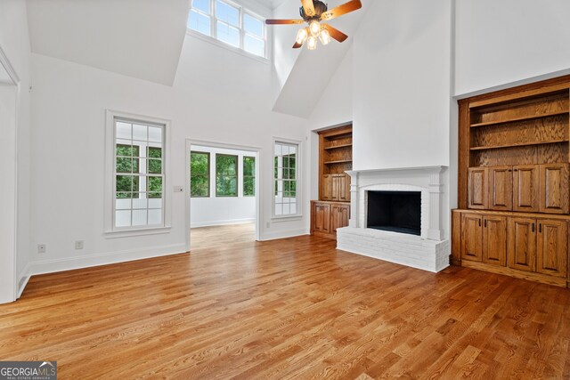 unfurnished living room featuring ceiling fan, a high ceiling, built in shelves, light hardwood / wood-style flooring, and a brick fireplace