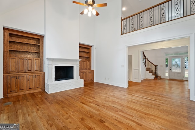 unfurnished living room featuring a fireplace, a high ceiling, light wood-type flooring, built in shelves, and ceiling fan