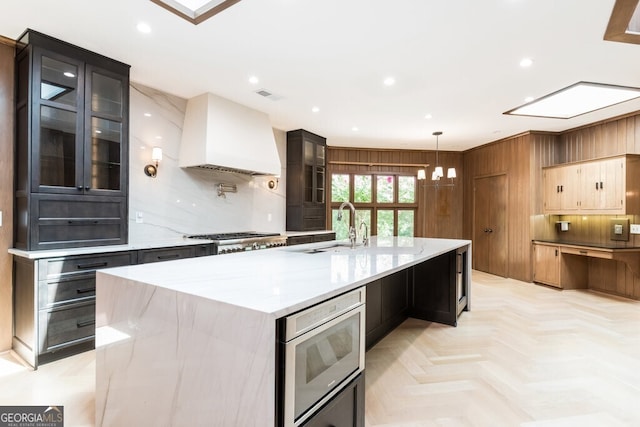 kitchen featuring pendant lighting, an island with sink, sink, light stone counters, and custom range hood