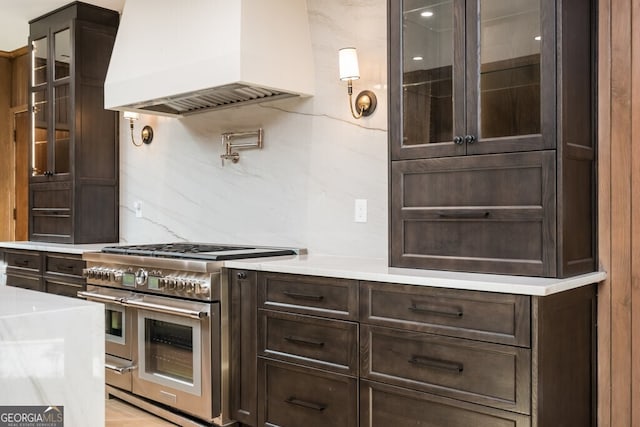 kitchen featuring double oven range, dark brown cabinets, premium range hood, backsplash, and light stone countertops