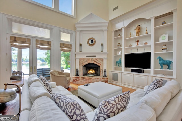 living room featuring a towering ceiling, wood-type flooring, french doors, built in features, and a brick fireplace