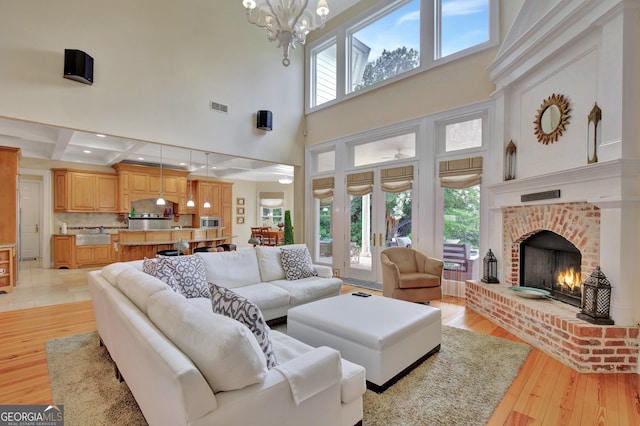 living room featuring beam ceiling, a notable chandelier, a brick fireplace, light wood-type flooring, and a towering ceiling