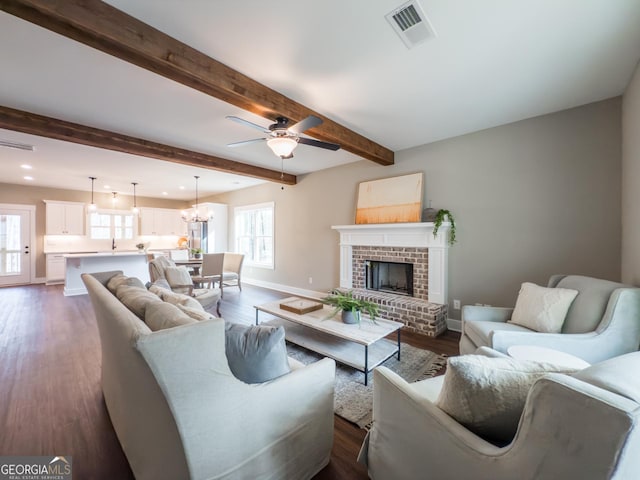 living room with dark wood-type flooring, ceiling fan with notable chandelier, a brick fireplace, and beamed ceiling