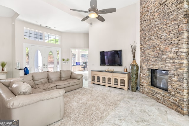 living room with french doors, ceiling fan, a stone fireplace, and a high ceiling