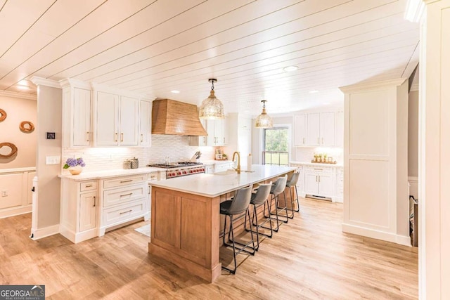 kitchen with premium range hood, hanging light fixtures, an island with sink, light hardwood / wood-style flooring, and backsplash