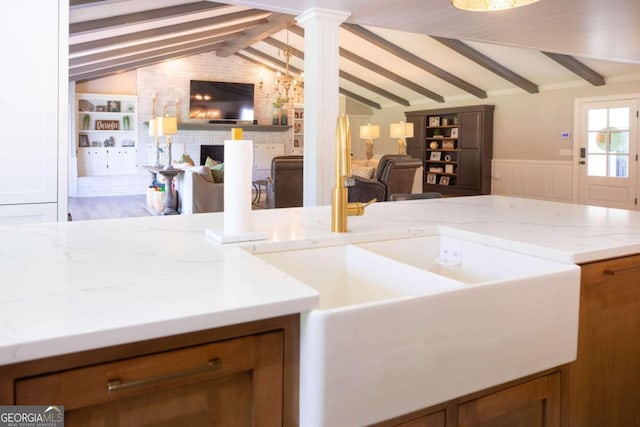 kitchen featuring built in features, lofted ceiling with beams, light stone counters, ornate columns, and a brick fireplace