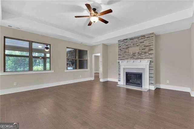 unfurnished living room with ceiling fan, a stone fireplace, dark wood-type flooring, and a tray ceiling