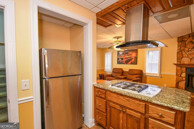 kitchen with a stone fireplace, island range hood, white gas stovetop, stainless steel fridge, and a drop ceiling