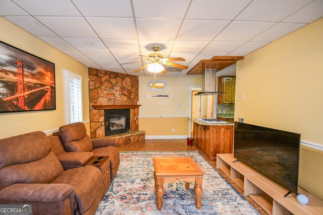 living room featuring ceiling fan, a stone fireplace, and a drop ceiling