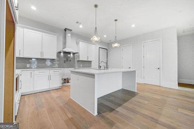 kitchen with white cabinets, hanging light fixtures, a kitchen island with sink, wall chimney exhaust hood, and light hardwood / wood-style flooring