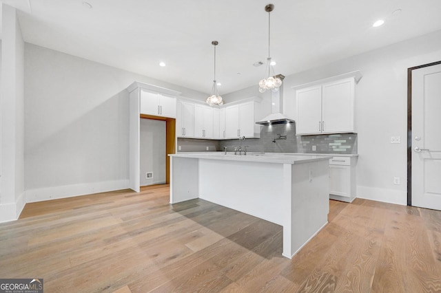 kitchen with pendant lighting, wall chimney range hood, white cabinetry, tasteful backsplash, and an island with sink