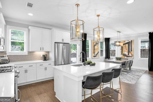 kitchen with crown molding, dark wood-type flooring, a kitchen island with sink, stainless steel fridge, and decorative light fixtures