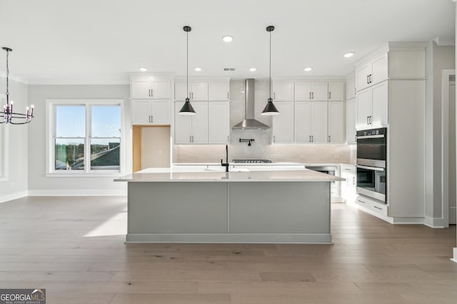 kitchen featuring wall chimney exhaust hood, stainless steel double oven, a kitchen island with sink, decorative light fixtures, and white cabinetry