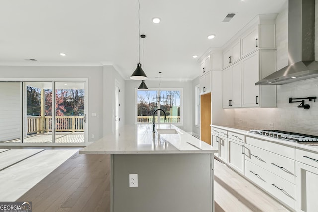 kitchen featuring white cabinetry, wall chimney exhaust hood, an island with sink, light hardwood / wood-style floors, and decorative light fixtures