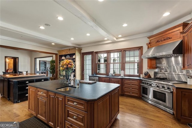 kitchen with appliances with stainless steel finishes, sink, a kitchen island with sink, wall chimney range hood, and beamed ceiling