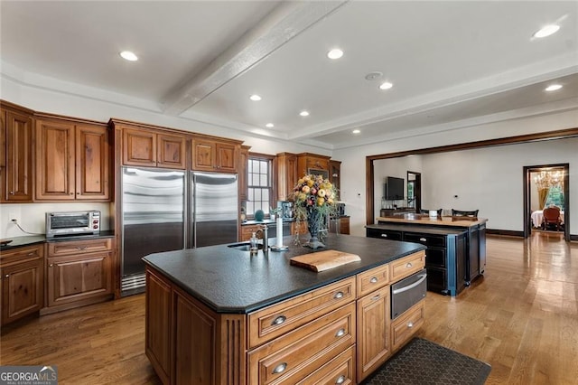 kitchen with light wood-type flooring, a kitchen island with sink, stainless steel built in fridge, and beamed ceiling