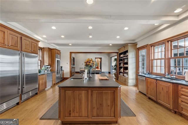 kitchen featuring appliances with stainless steel finishes, beamed ceiling, an island with sink, sink, and light wood-type flooring