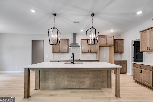 kitchen featuring a kitchen island with sink, wall chimney exhaust hood, sink, decorative light fixtures, and light wood-type flooring