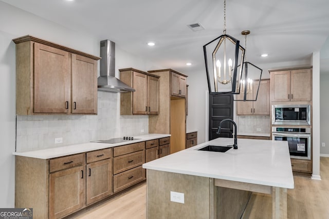 kitchen with wall chimney range hood, sink, stainless steel appliances, an inviting chandelier, and a center island with sink