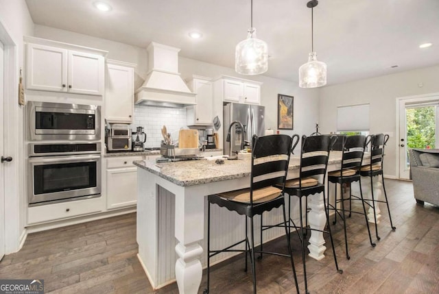 kitchen featuring stainless steel appliances, custom range hood, white cabinets, and a center island with sink