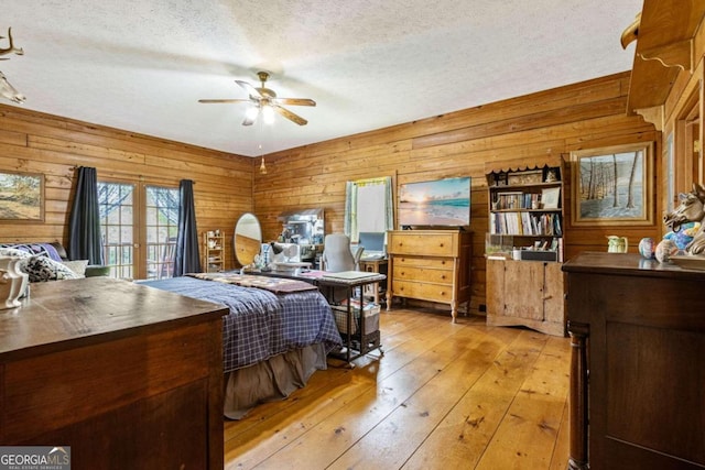 bedroom featuring ceiling fan, a textured ceiling, light hardwood / wood-style flooring, wooden walls, and french doors