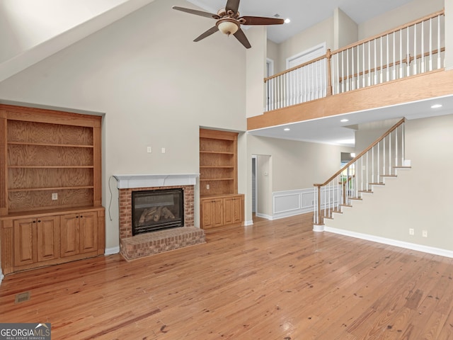 unfurnished living room with a brick fireplace, a towering ceiling, stairway, light wood-type flooring, and a ceiling fan