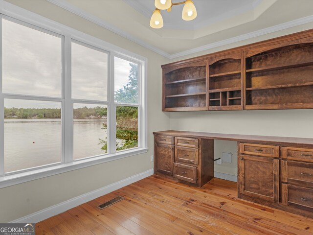 unfurnished office with crown molding, a water view, a tray ceiling, built in desk, and light wood-type flooring