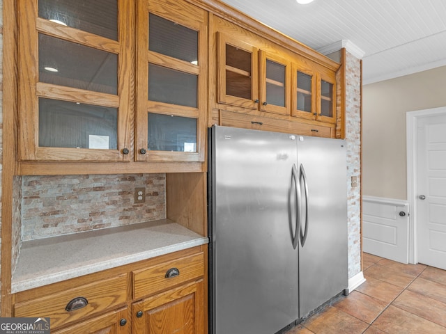 kitchen featuring brown cabinetry, light tile patterned floors, freestanding refrigerator, glass insert cabinets, and ornamental molding