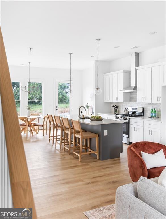 kitchen with stainless steel range with gas cooktop, an island with sink, decorative backsplash, white cabinetry, and wall chimney exhaust hood