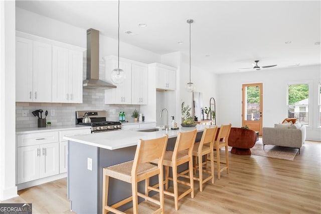 kitchen featuring wall chimney range hood, an island with sink, stainless steel gas range oven, sink, and light hardwood / wood-style floors