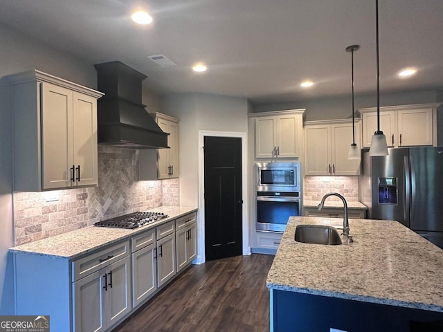 kitchen with sink, appliances with stainless steel finishes, an island with sink, custom range hood, and pendant lighting