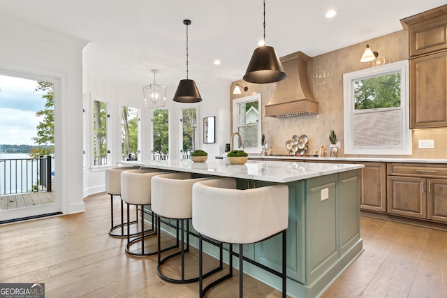 kitchen with light stone counters, custom exhaust hood, a large island with sink, backsplash, and light wood-type flooring