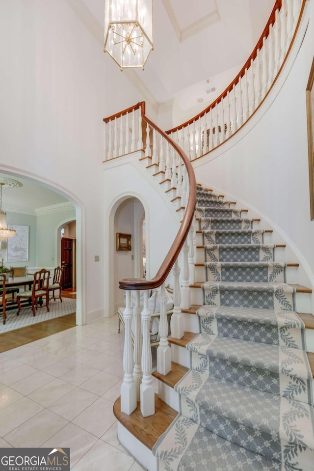 stairs with arched walkways, crown molding, a towering ceiling, an inviting chandelier, and tile patterned flooring