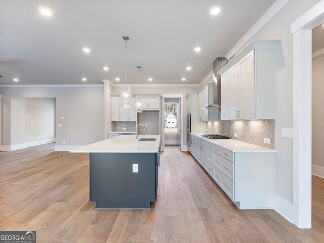 kitchen featuring white cabinetry, wall chimney range hood, light hardwood / wood-style flooring, pendant lighting, and a kitchen island with sink