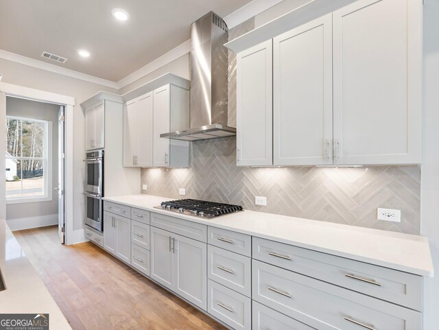 kitchen with wall chimney range hood, light hardwood / wood-style flooring, backsplash, appliances with stainless steel finishes, and ornamental molding