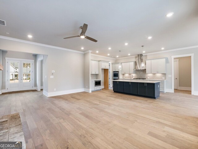 kitchen with pendant lighting, a kitchen island with sink, white cabinets, wall chimney range hood, and light hardwood / wood-style flooring