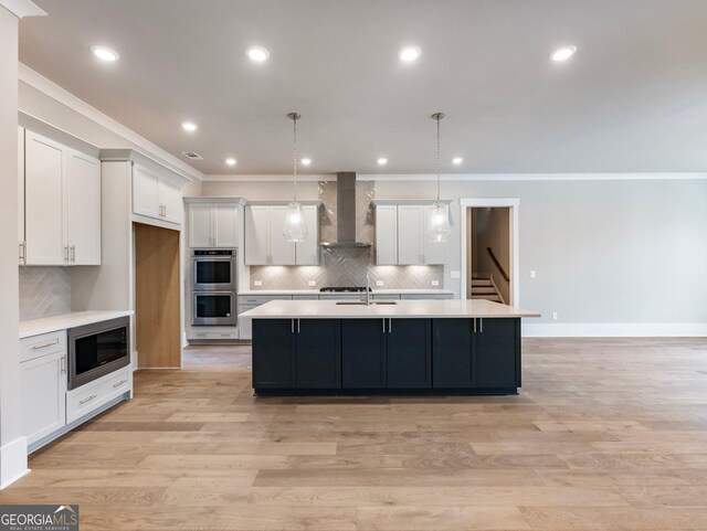 kitchen with appliances with stainless steel finishes, wall chimney range hood, white cabinetry, hanging light fixtures, and an island with sink