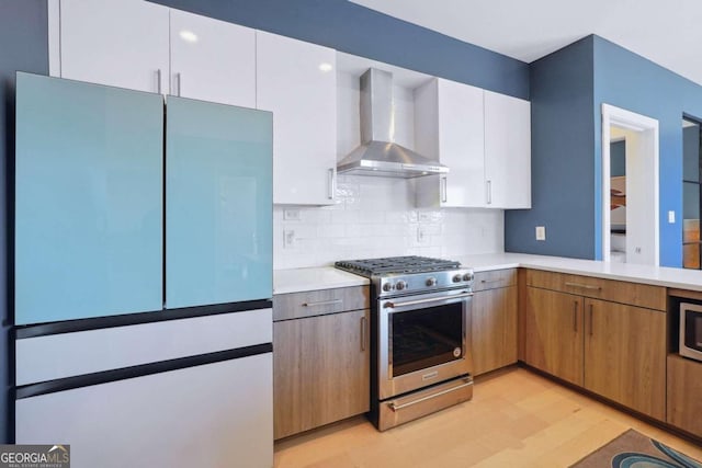 kitchen featuring white cabinetry, light wood-type flooring, wall chimney range hood, backsplash, and appliances with stainless steel finishes