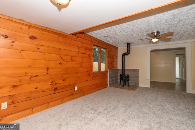 unfurnished living room with a wood stove, carpet flooring, ceiling fan, and wooden walls