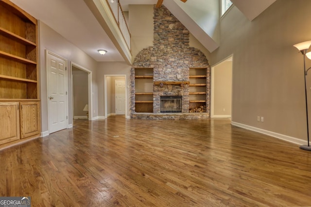 unfurnished living room with built in features, a towering ceiling, a stone fireplace, wood finished floors, and baseboards