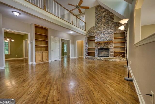 unfurnished living room featuring built in features, a stone fireplace, ceiling fan, wood-type flooring, and a towering ceiling