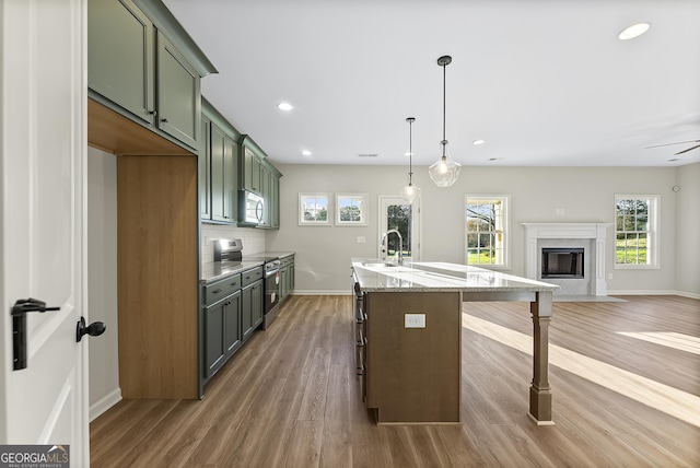 kitchen featuring light stone counters, electric stove, a center island with sink, green cabinetry, and hanging light fixtures