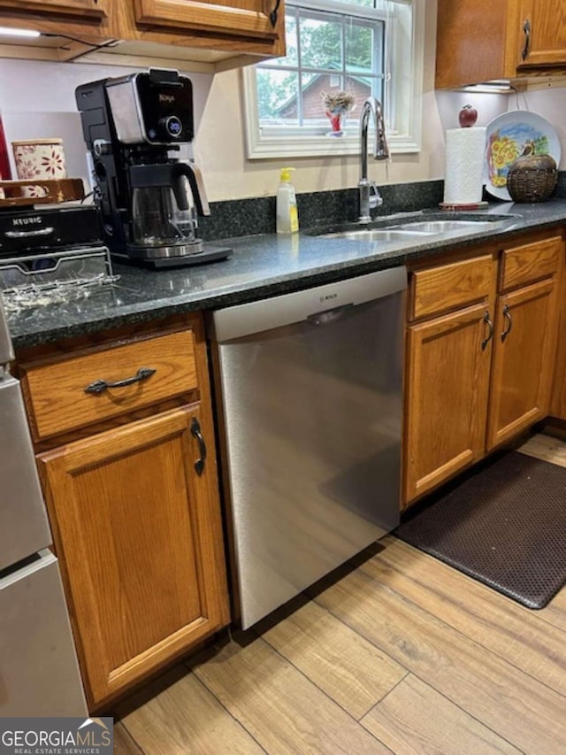 kitchen with brown cabinets, dark stone countertops, light wood-type flooring, stainless steel dishwasher, and a sink