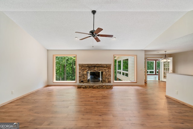 unfurnished living room featuring light wood-type flooring, ceiling fan with notable chandelier, a textured ceiling, a stone fireplace, and vaulted ceiling