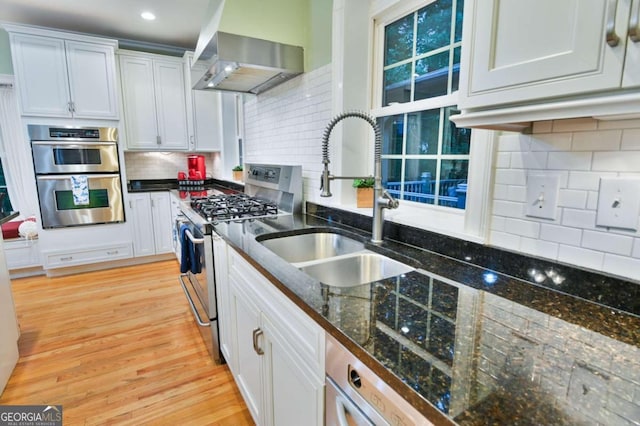 kitchen featuring wall chimney range hood, decorative backsplash, light hardwood / wood-style flooring, white cabinetry, and stainless steel appliances