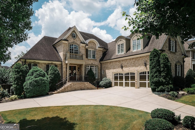 french country style house with driveway, an attached garage, roof with shingles, and brick siding